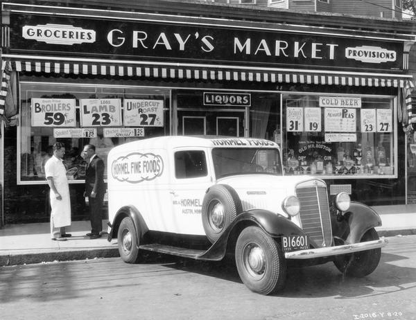 Two men standing outside Gray's Market behind an International C-1 delivery truck owned by Hormel Fine Foods. One man is wearing an apron and holding a canned ham. Truck featured a 125-inch wheel base and panel body.