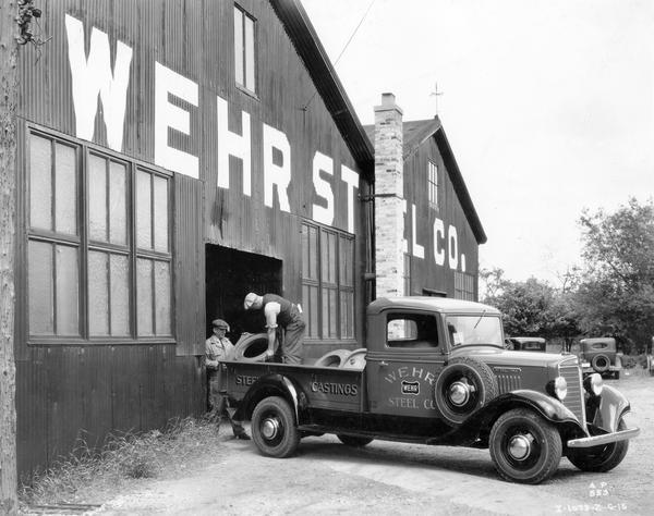 Two workers loading large steel castings into the back of an International C-15 truck outside a Wehr Steel Company building.