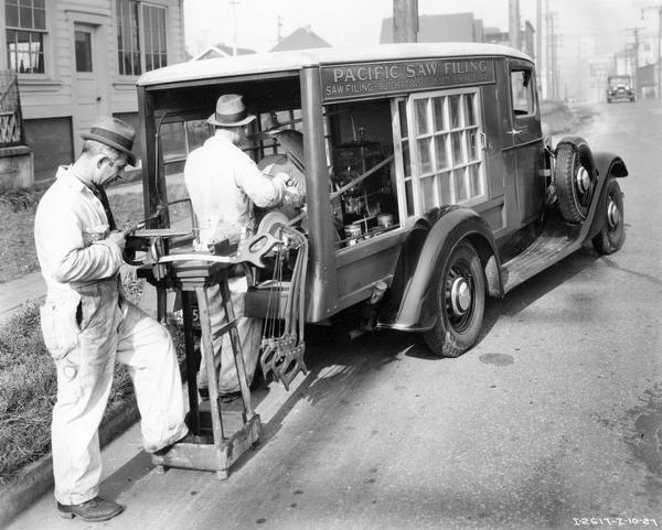 Two men sharpening tools from the back of an International C-5 truck equipped with a special body and grinding equipment. The truck was owned by Pacific Saw Filing. The men are, (L to R): John Ciffone and William Richard. The filing business was located at the corner of Keefer and Vernon Drive in Vancouver, British Columbia.