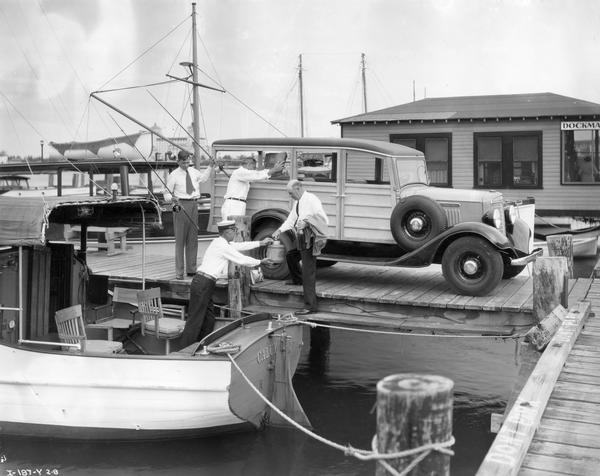 Three men on a dock unloading gear from an International C-1 wood-paneled ("woody") station wagon for a fishing trip. One man is standing in a fishing boat called the "Calumet." A dockmaster's building is in the background.