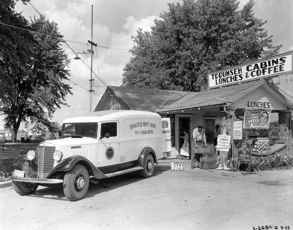 Milkman unloading crates from an International C-15 truck in front of "Tecumseh Cabins Lunches, & Coffee". The front of the restaurant and store features several signs, including advertisements for the Sunday night Eddie Cantor program on the Columbia Network. The truck featured a special 9-foot body and was owned by Springfield Purity Dairy.