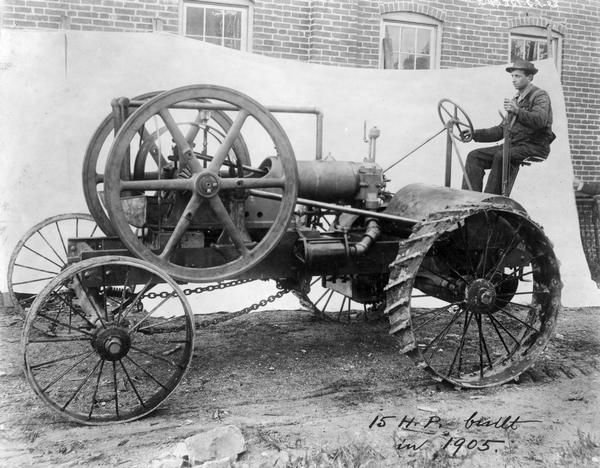 Man posing in the seat of a 15 HP tractor "built in 1905." A white backdrop is behind the tractor, and a brick building with windows is behind the backdrop.