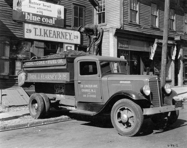 Man standing in the bed of an International coal delivery truck owned by Thos. J. Kearney Jr., Inc. The man is shoveling coal and the truck is backed up to the front of Kearney's place of business. An advertisement on the building reads "Better heat, less attention with 'blue coal'."