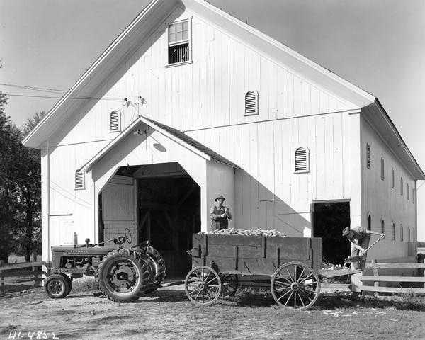 Delmer Miller is standing in a farm wagon while farm hand, Gus Setser, is shoveling corn from the back end. The wagon is hitched to a Farmall H tractor and is parked in front of Mr. Miller's barn. The tractor was equipped with Firestone ground grip tires.