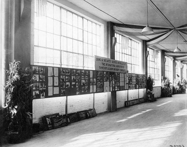 Pictures and diagrams mounted on a wall in a "new building" at International Harvester's Deering Works. The display was part of an industrial exhibit and depicts the steps the company had taken to improve employee safety inside its factories. A banner above exhibit reads: "Some of the safety devices installed by the International Harvester Co. to prevent accident and injury to employees."