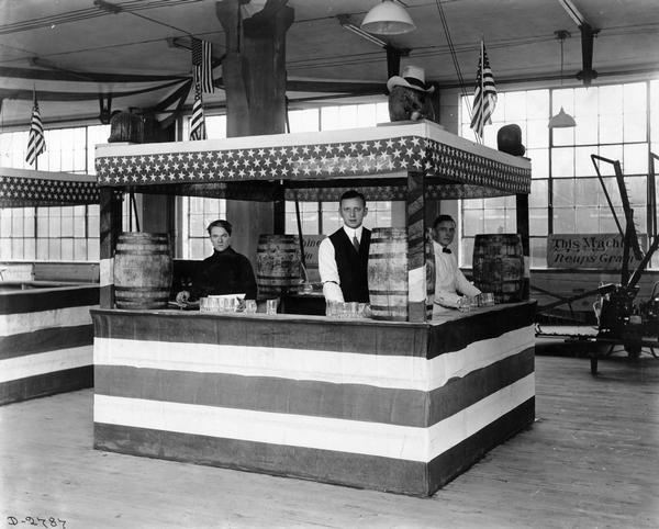 Three workers with barrels of drinks inside a refreshment booth decorated with stars and stripes. The men were preparing to serve drinks at an International Harvester industrial exhibit, most likely taking place at the company's Deering Works. A reaper is on display in the background.