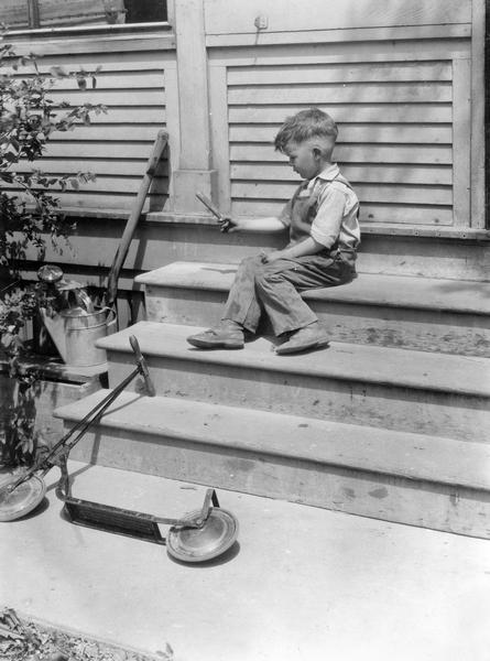 Staged scene of young boy about to light large firecracker. He is sitting on steps, and a push scooter is lying on its side on the sidewalk. This photograph was taken for International Harvester's Agricultural Extension Department at the company's experimental farm. It was used to warn families of potential farm hazards.