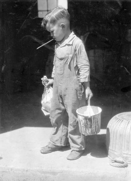 Young farm boy holding a paint bucket and paper bag and has a pencil in his mouth. This photograph was taken for International Harvester's Agricultural Extension Department at the company's experimental farm. It was used to warn families of potential farm hazards. The original caption reads: "Teach children to keep things out of mouth."