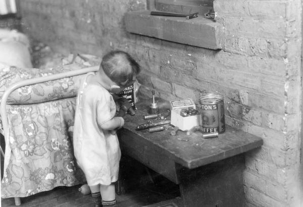 Staged scene of a toddler playing with shot gun shells and a hammer. This photograph was taken for International Harvester's Agricultural Extension Department. It was used to warn families of potential farm hazards. The original caption reads: "Youngster playing with shot gun shells. They explode easily."