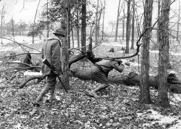 Staged scene of two hunters in the woods with guns. One man is walking under a branch with his  rifle pointed behind him. This photograph was taken for International Harvester's Agricultural Extension Department. It was used to warn families of potential farm hazards. The original caption reads: "Walking single file and crawling under limbs and fences is dangerous with a loaded gun while hunting."