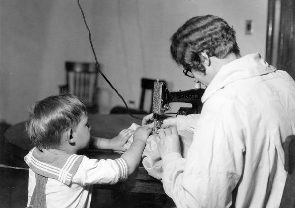 Staged scene of a woman sewing with a young child attempting to help. This photograph was taken for International Harvester's Agricultural Extension Department at the company's experimental farm. It was used to warn families of potential farm hazards. The original caption warns of children "getting fingers too close to sewing machine needle."