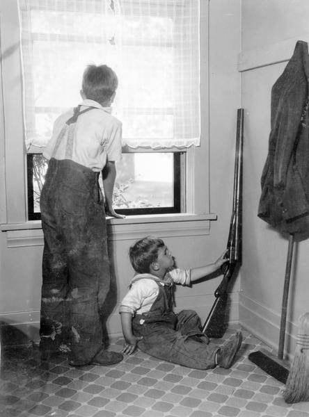 Staged scene of two young boys near a window with one reaching for a shotgun. This photograph was taken for International Harvester's Agricultural Extension Department at the company's experimental farm. It was used to warn families of potential farm hazards. The original caption warns of "having guns within reach of children."
