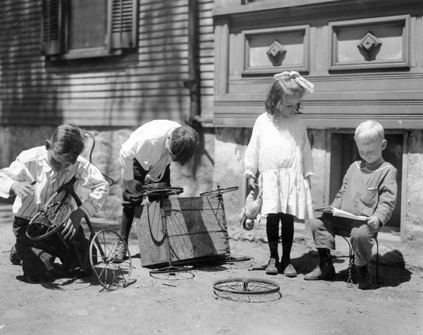 Three young boys and one girl at play near a house. Two boys are fixing the wheels on a tricycle and wagon. The girl, holding a doll, is looking on and the third boy is sitting reading a book.