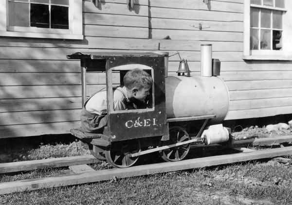 Kenneth Chipman riding a toy railroad engine on the farm of W.W. Chipman. Worked by footpower and with a working whistle bell, the toy engine follows tracks set up across the farm yard. The engine was made by a local miner and old C&EI railroad employee.