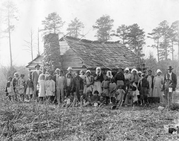 Group portrait of African American children and adults posing in front of a run-down building with a stone chimney — possibly a rural school house.