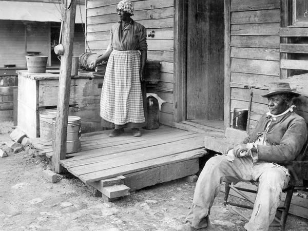 African-American man and woman on the front porch of a rural home. The unidentified woman is retrieving water from a well, and the man, J.R. Dean, is sitting in broken chair.