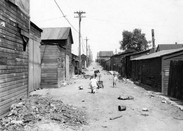 Three young children playing with a wagon in a squalid alley between rows of impoverished houses. Trash and rubble litter the alley.