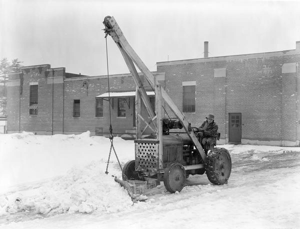 Worker plowing snow with a McCormick-Deering industrial tractor equipped with a crane and blade.