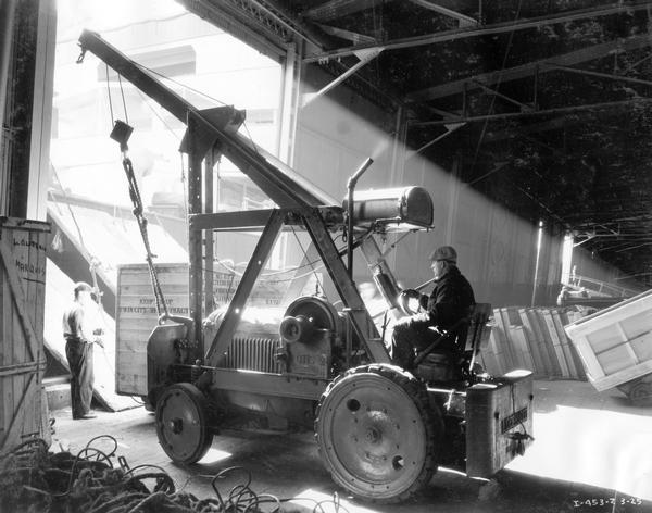 Dock worker using an International Model 20 tractor equipped with a Lidgerwood crane to load cargo onto a ship.