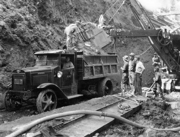 Workers are looking on as a Bucyrus Erie excavating crane is loading dirt into the bed of an International heavy-duty dump truck.