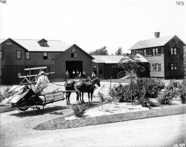 Farmer on a horse-drawn McCormick binder outside a large farmstead as men, boys, and a dog are looking on. One boy is on a bicycle.