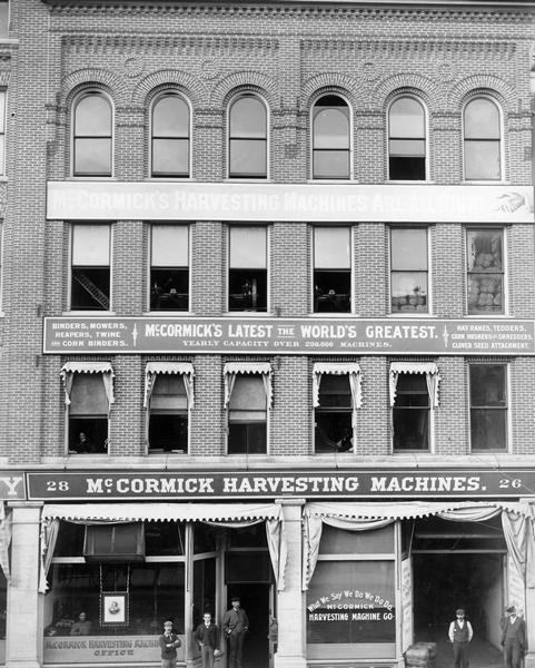 View from across street of the three-story facade of a large brick building. A group of employees are posing outdoors in front of the McCormick Harvesting Machine Company dealership. A woman is standing behind the plate glass window next to a portrait of Cyrus Hall McCormick. A slogan on the storefront window reads: "What we say we do we do do." Implements and stacks of twine can be seen through the upper story windows. The general agent was Mr. Frazier.
