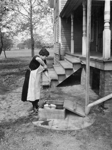 Miss Neary drawing a bucket of water from a well next to a farmhouse. The original caption reads: "Drawing water from well. Miss Neary posing. Mr. Hyde photographer."