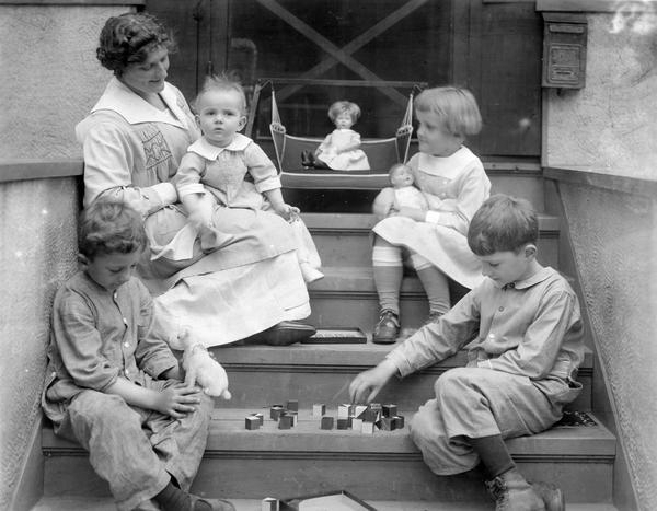 Mother and her four children playing on the steps of a house. The children are playing with blocks, dolls and a stuffed animal.
