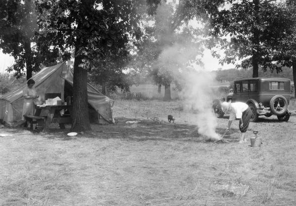 Man tending to a campfire while a woman is preparing a meal on a picnic table by a tent. A car is parked next to the campsite and a dog is sniffing at a tent stake. The photograph was taken for International Harvester's Agricultural Extension Department to demonstrate potential "farm hazards." The original caption reads: "Carelessness--leaving camp fire burning may start a fire."