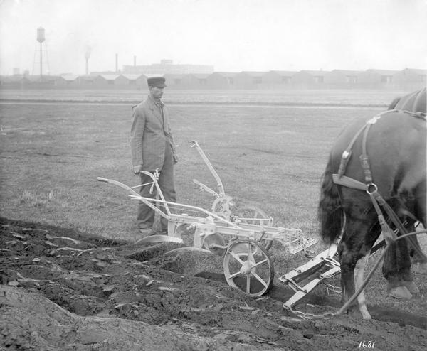 Man operating an Oliver no. 36 horse-drawn walking plow in a field with factory buildings in the background. The photograph was taken for, or compiled by International Harvester's Agricultural Extension Department.