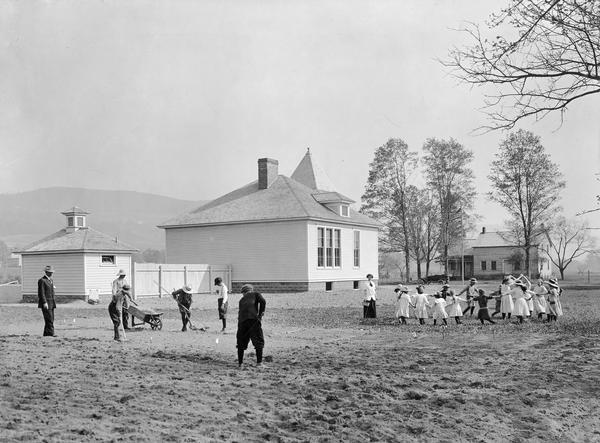 Boys working in a garden while smaller children dance in a circle on the grounds of Middlefield No. 2 Consolidated School in Ostego County. Teachers are looking on. The schoolhouse and other buildings are in the background.