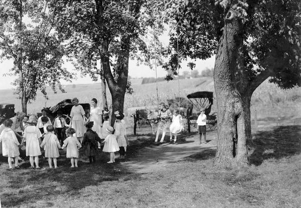 Children and a few adults (possibly teachers) hold hands in a circle while other children stand by or swing under a tree in a rural setting. Horse-drawn carriages are parked in the background. The photograph was taken for International Harvester's Agricultural Extension Department and is part of an album devoted to "schools."