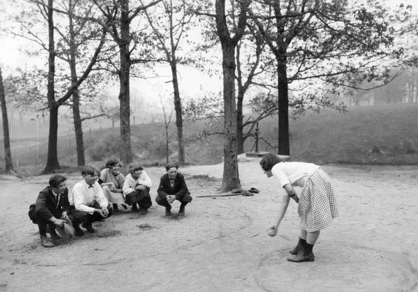 Country school children playing "Baby in the Hole." There is a hand-pump in the background under trees.