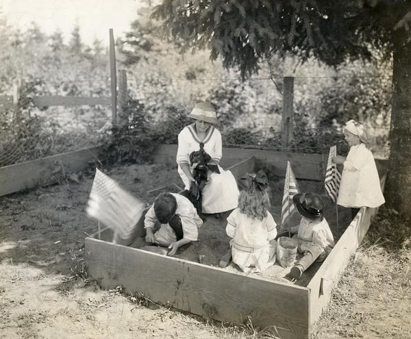 Children playing in a sand box, or "sand garden" at the Woodlawn School garden. American flags are planted in the sand and a teacher or older child is holding a dog.