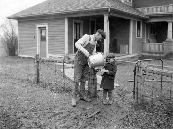 Farmer Hal Ament pouring milk from a pail into a glass for his young son as they stand at the gate of their farmhouse.