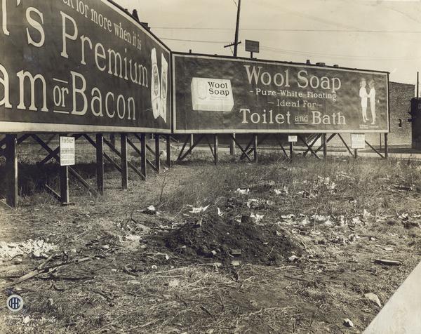 Vacant city lot cluttered with litter and advertising billboards for Premium Ham and Bacon and Wool Soap. The department likely used the photograph to show a site that could be cleared and used for a city garden.