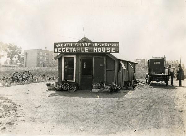 Small vegetable store known as the North Shore (Home Grown) Vegetable House, along the north shore of Lake Michigan. To the right of the store two men are talking next to a truck. Original caption reads: "This man raised his vegetables and marketed them on the ground by building a small store for the purpose."