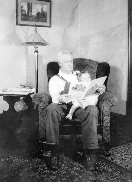 Mr. Wolfe sitting in a chair reading <i>Successful Farming</i> magazine to a young child on his lap.