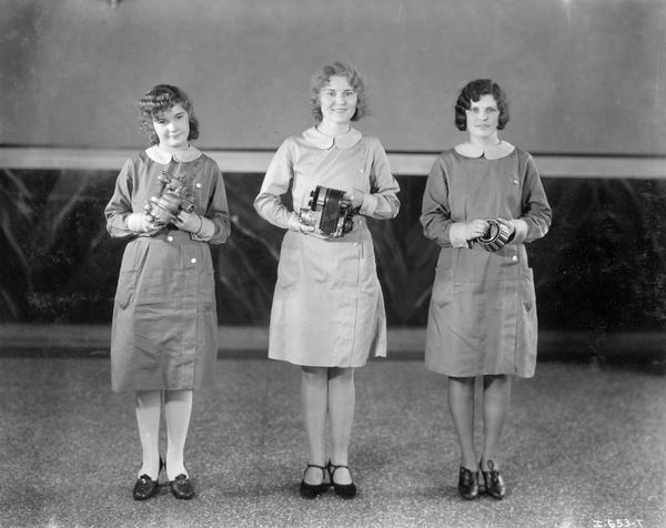 Three female workers standing and holding what appear to be a carburetor, magneto and roller bearing at International Harvester's West Pullman Works.