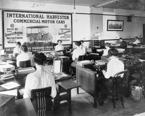 Women and men at work inside an office. The women are using typewriters. A large advertisement for International motor trucks is on one wall.