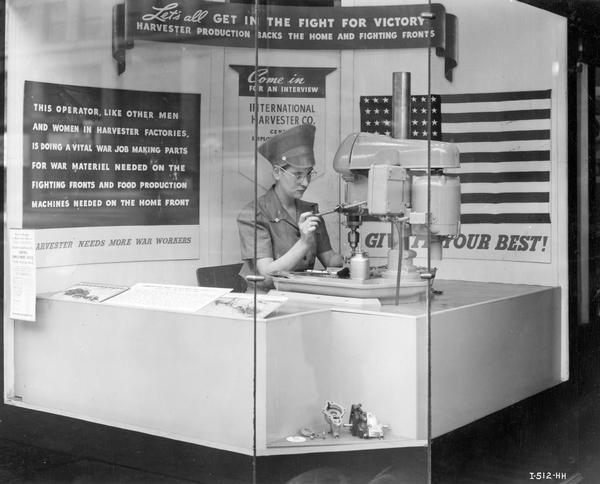 Female worker using a drill press in a window display at International Harvester's employment office in the company's Michigan Avenue annex. The display celebrates war work. A sign in the display reads: "This operator, like other men and women in Harvester factories, is doing a vital war job making parts for war materiel needed on the fighting fronts and food production machines needed on the home front."