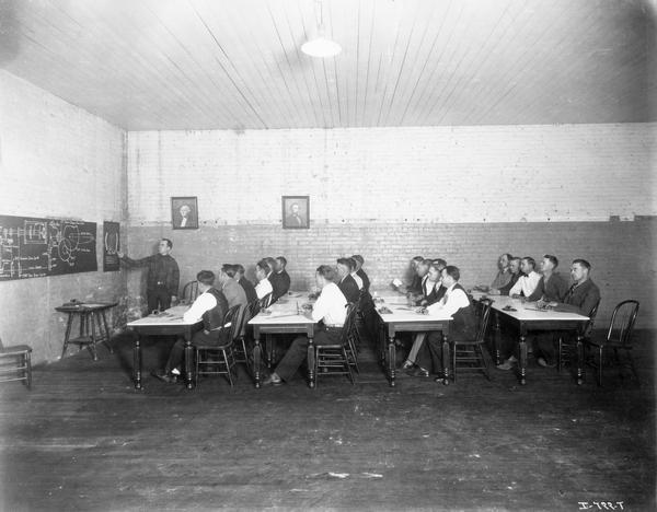 Employees(?) learning about a chain drive gear at the St. Paul Flax and Twine Mill. Portraits of George Washington and Abraham Lincoln and an advertising poster for the "big ball" of twine are hanging on the brick walls. The factory was owned and operated by an International Harvester subsidiary called the International Flax Twine Company.