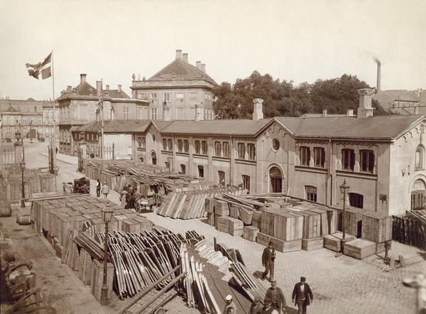 Elevated view of stockyard outside a McCormick Harvesting Machine Company dealership in Copenhagen, Denmark. The yard is filled with boxes containing McCormick farm machines and implements and several people are milling about. The Amalieuborg Castle is in the background.