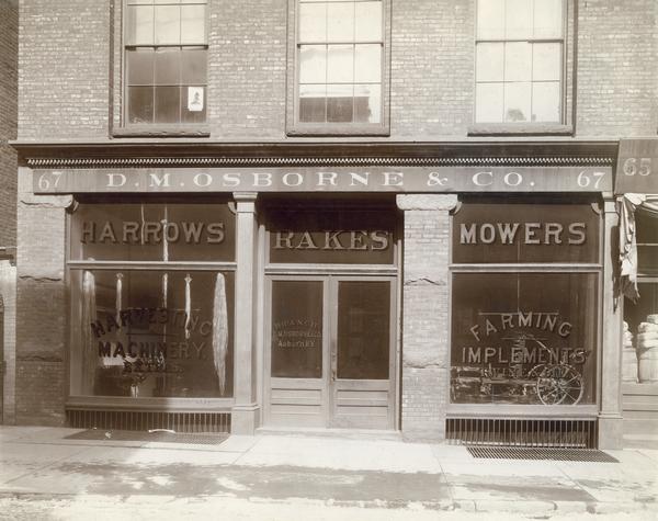 Storefront of D.M. Osborne & Company dealership building. The words "harrows," "rakes," and "mowers" are painted on the windows. Strands of binder twine are hanging in one window and a mower is displayed in the other. Osborne was a farm implement manufacturer that was purchased by the newly formed International Harvester Company in 1903.