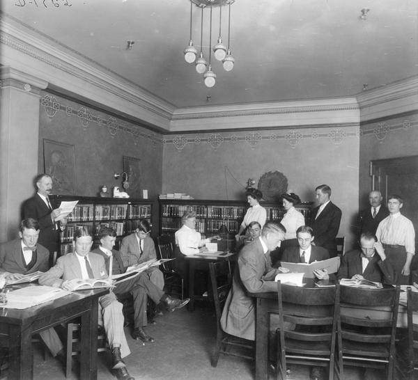 Office(?) workers reading in the library at the Deering Works Club. In the background the librarian (seated at desk) appears to be checking books in and out.