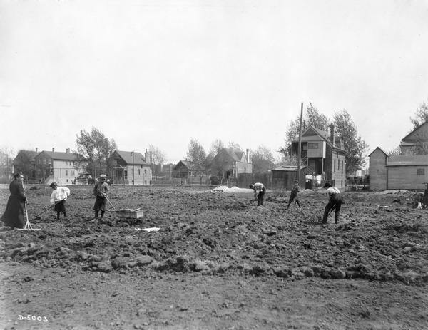 Men, women and children dig and rake the soil at the Deering Works employee gardens. The gardens appear to be located in a residential neighborhood.