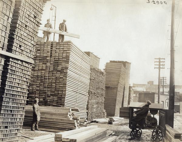 Four employees hoisting planks of wood onto large stacks with a portable electric winch in the stock yard at International Harvester's Deering Works.