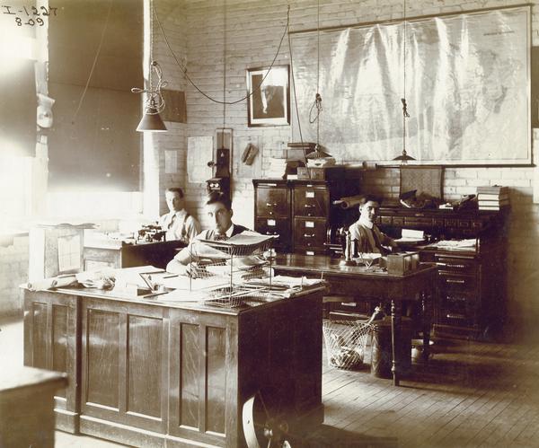 Three office workers at their desks at an International Harvester office in Canada(?). A map of Canada and a portrait of William Deering are hanging on the wall behind them.