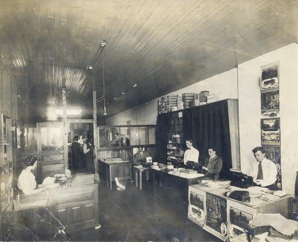 Men and women at work in the office of an International Harvester branch house(?). Advertising posters for Columbus wagons, Bluebell cream separators and International Harvester hay presses adorn a wall and a desk.