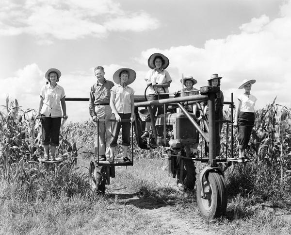 Six women and a man on a McCormick-Deering Farmall Cub tractor modified for de-tasseling work. The unit was built with a special mounting frame by Knudsen Implement Co., Manning, IA. The tractor was owned and operated by Gruhn Hybrid Corn of Manilla, IA.